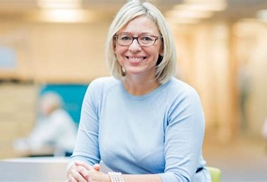 PVC Students and Education, Claire Morgan sits in a library. She wears a blue blouse, glasses and a pearl bracelet. She is smiling and has her hands clasped.