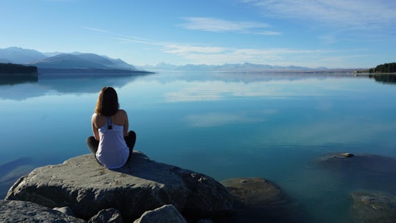 A decorative image. A female student sits cross legged on a grey rock with her back to the camera. She stares out onto a peaceful blue lake bordered with rugged mountains and forestry (New Zealand).