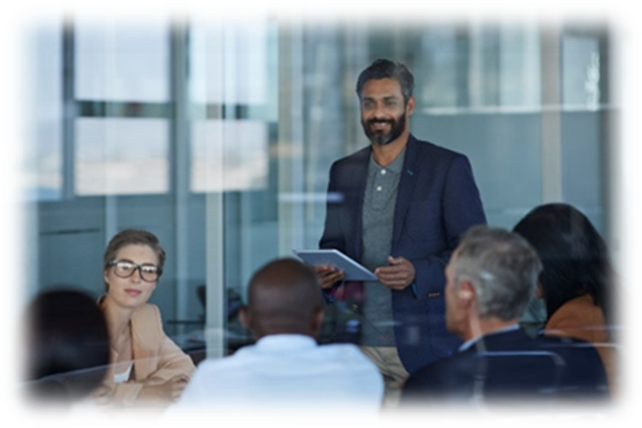 A decorative image meant to represent a programme director talking with his programme team. He wears a blue blazer and stands with a wad of paper documents in his hands, smiling at his team of 5 staff who are gathered around a table in a circle.