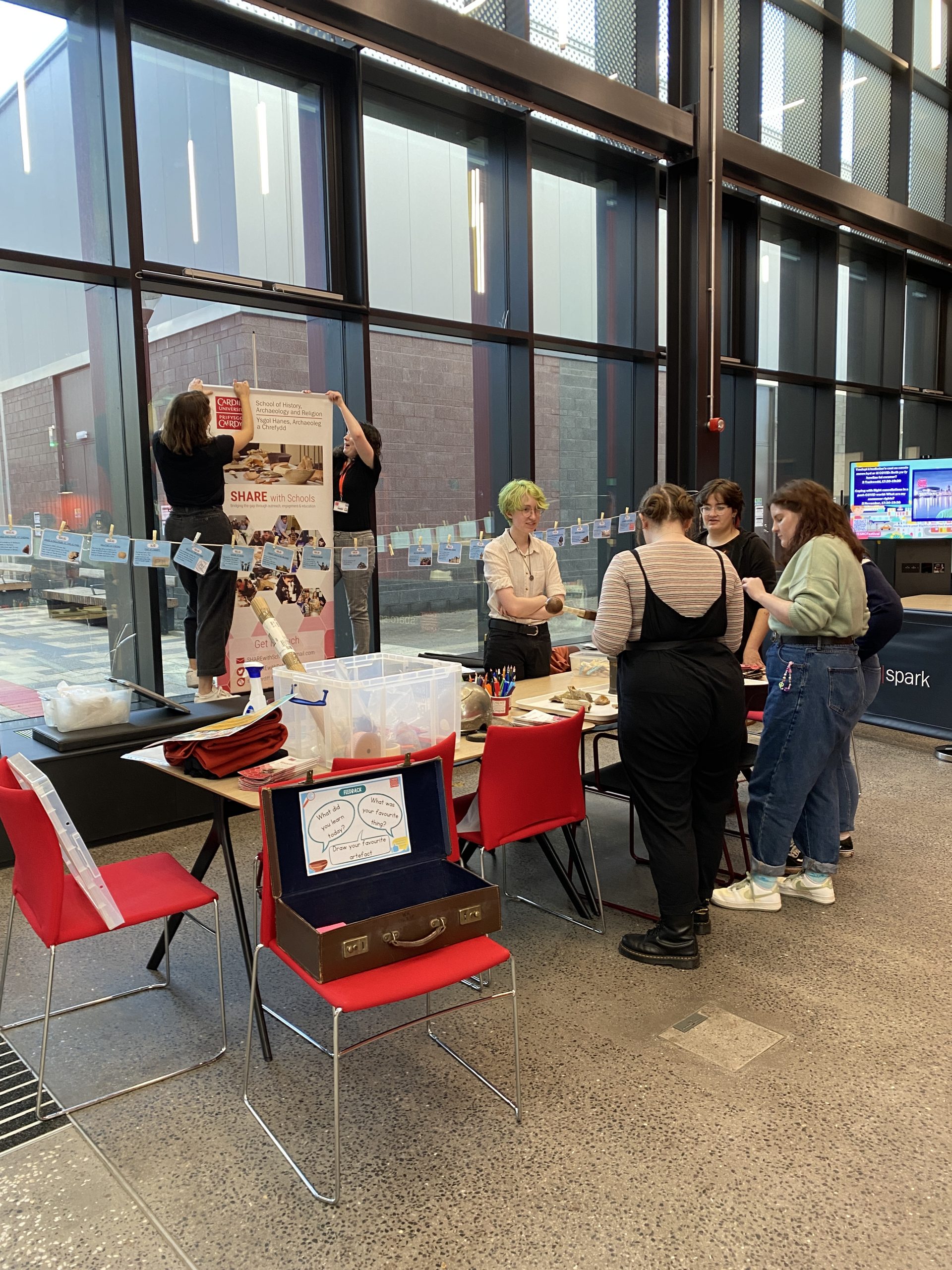 SHARE with Schools team setting up for the event. A group of 4 volunteers and a postgraduate coordinator are setting up handling objects on the table in the foreground. Two coordinators are hanging up a SHARE with Schools banner in the background.