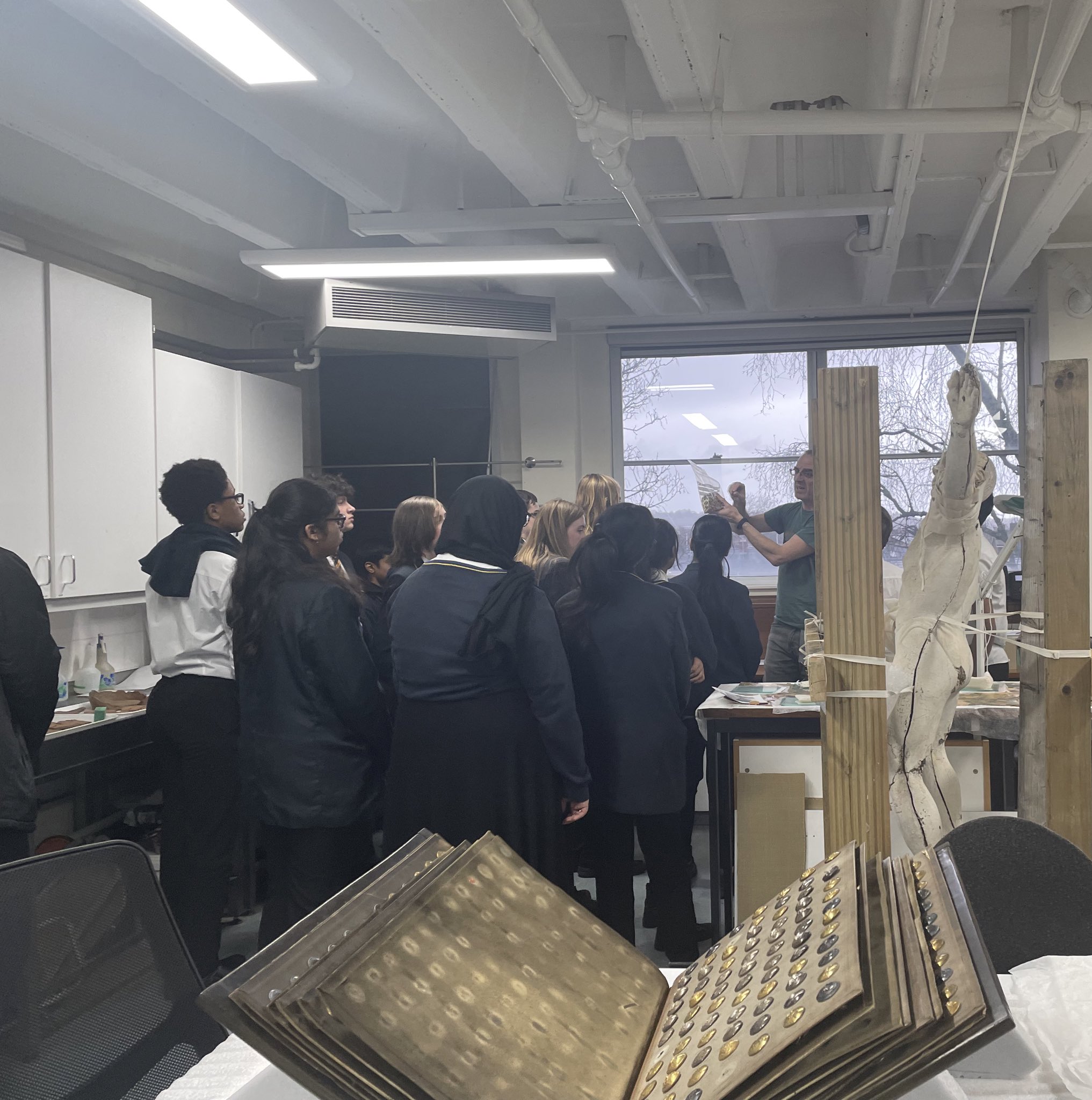 A group of young people gather around, listening to an academic member of staff holding a bag of cotton swabs as he explains their use. A book of badges and a model of a human are in the foreground in the conservation lab.