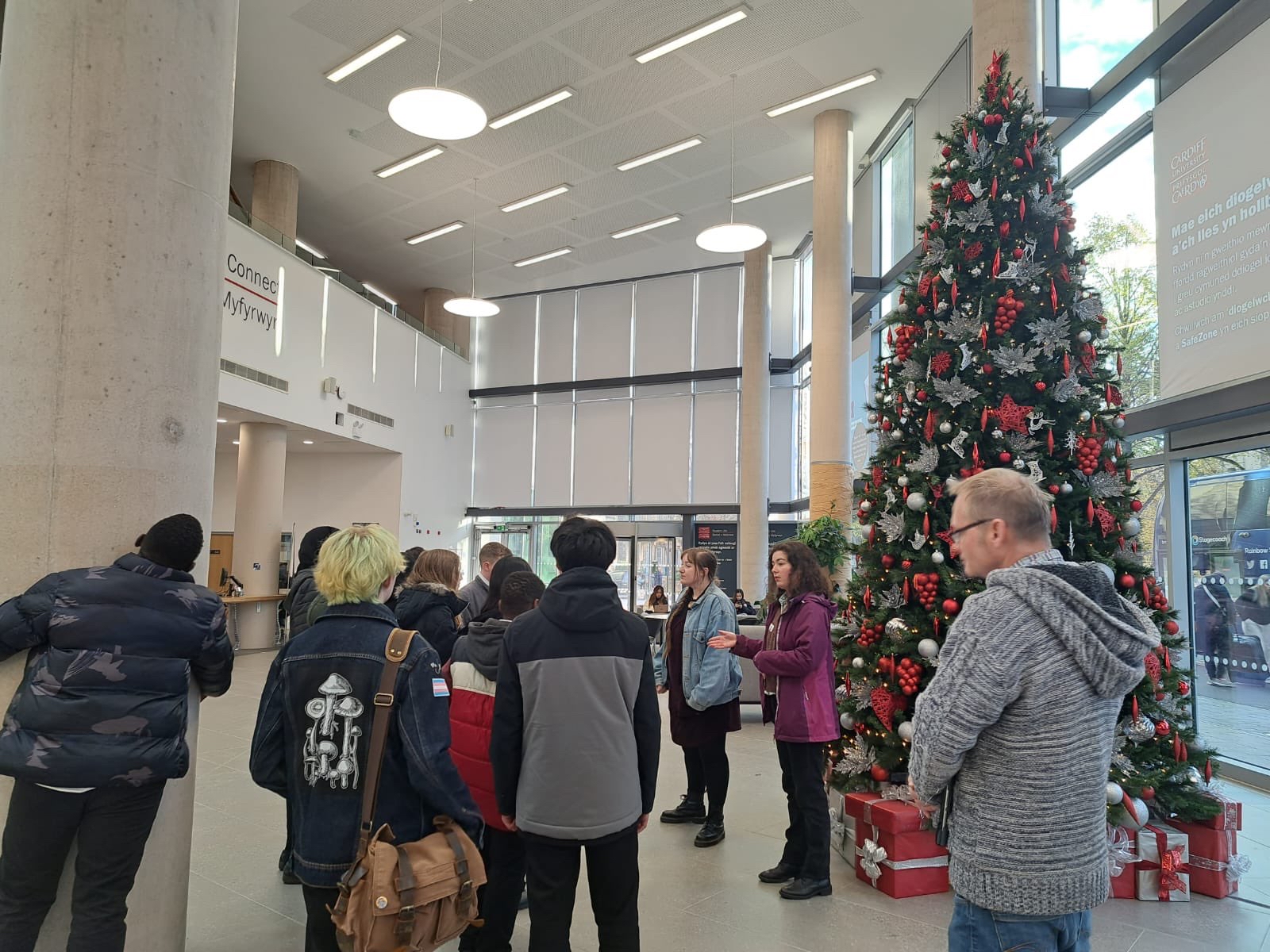 2 SHARE with Schools coordinators standing in front of a large Christmas tree speaking to a group of secondary school pupils. 