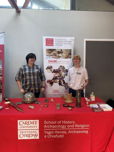 Two university students stood behind a table. On the table is a series of archaeological artefacts such as pottery, a roman sword and miners lamp.