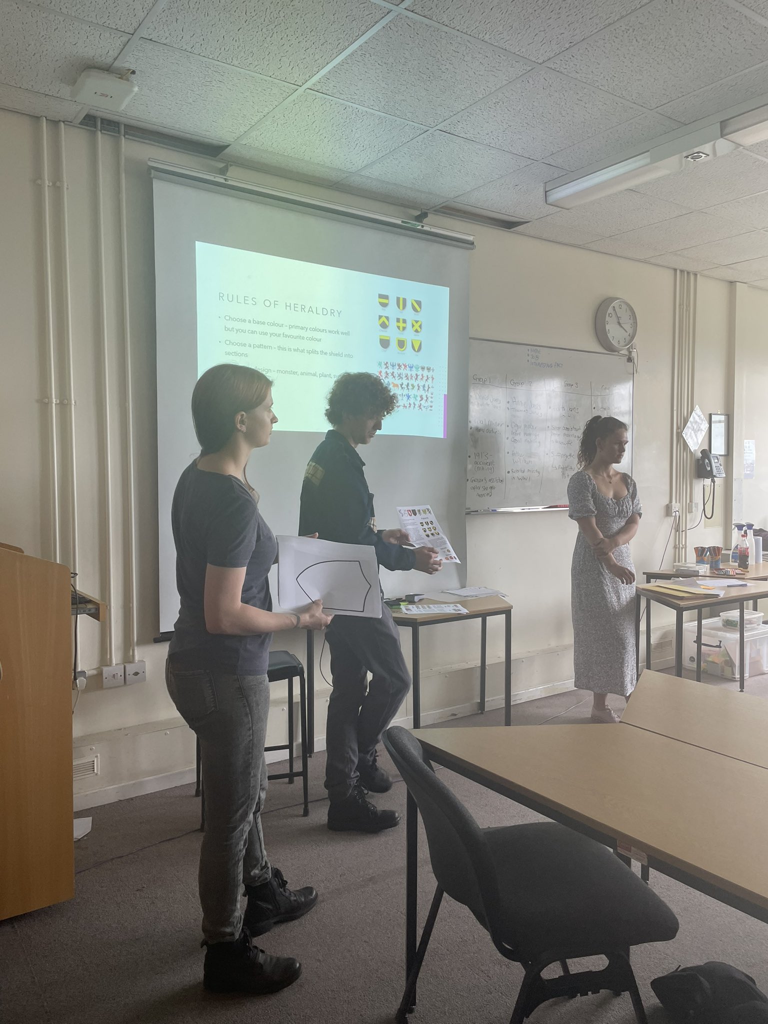 Three people stand at the front of a seminar room, presenting on the rules of heraldry