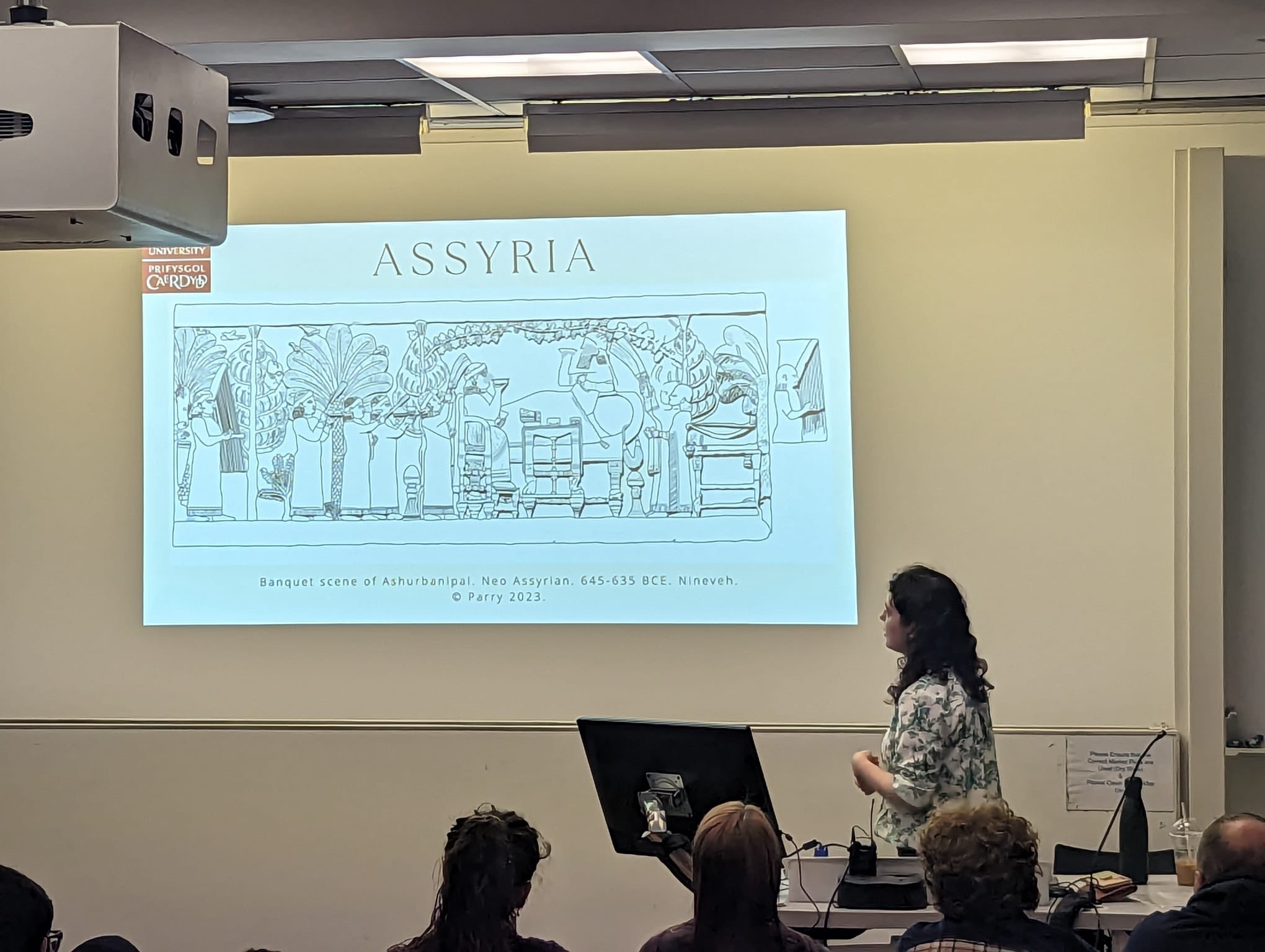 A woman stands at the front of a lecture hall. The slide depicts a line drawing go Ashurbanipal's banquet relief with the title 'Assyria'