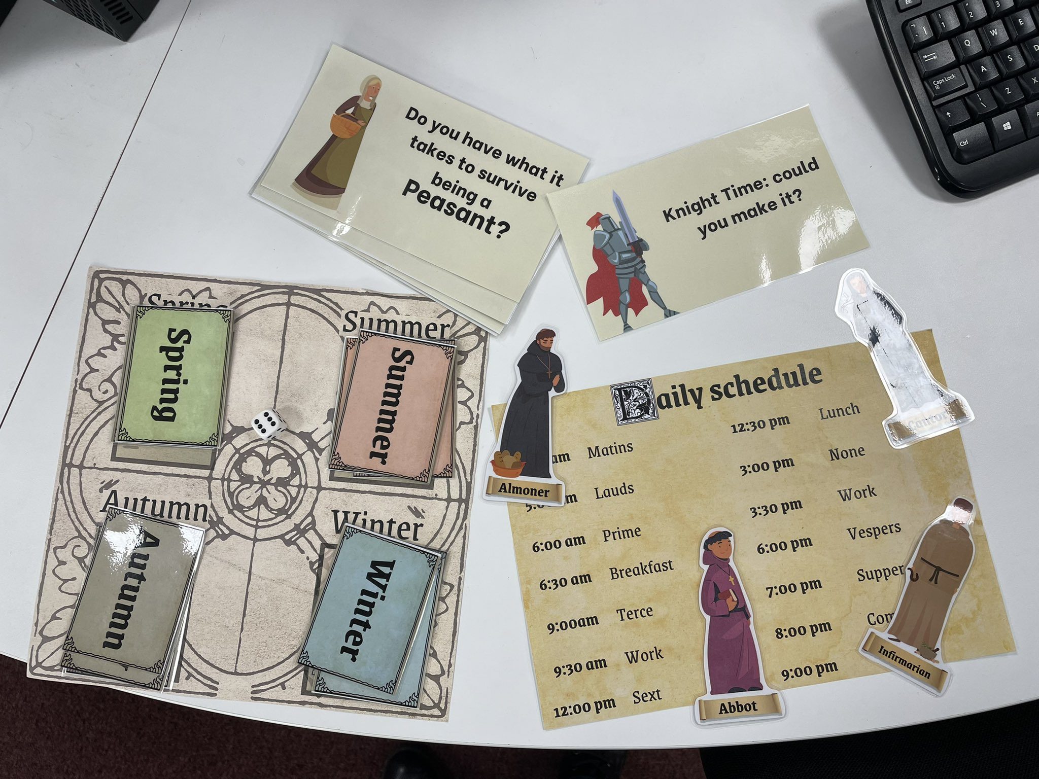 Medieval workshop resources on a table, showing cards from a peasant role-playing game, quiz cards about life as a monk and knight, and a copy of a medieval monk's daily schedule.