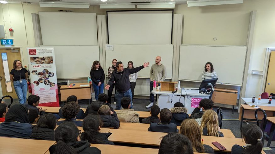 Young people sat in a lecture theatre facing the front where students and staff from the university stand speaking to the room.