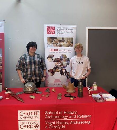 Two university students stood behind a table. On the table is a series of archaeological artefacts such as pottery, a roman sword and miners lamp.