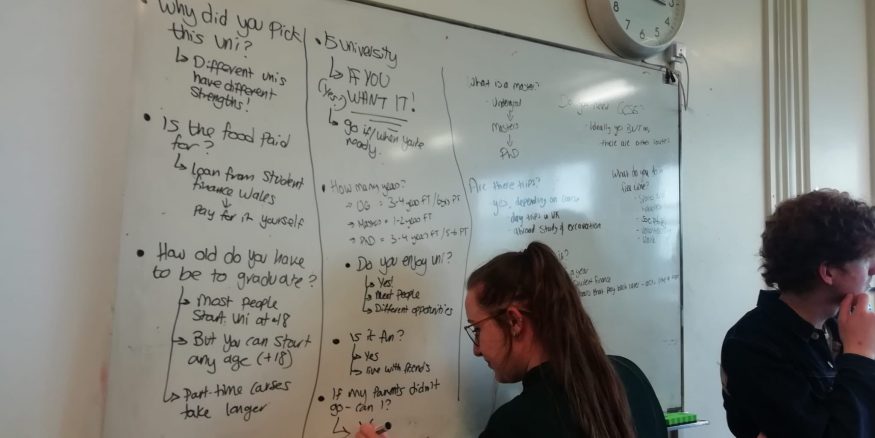 A woman in a green shirt writes on a whiteboard. On the whiteboard are a list of questions with answers about university,