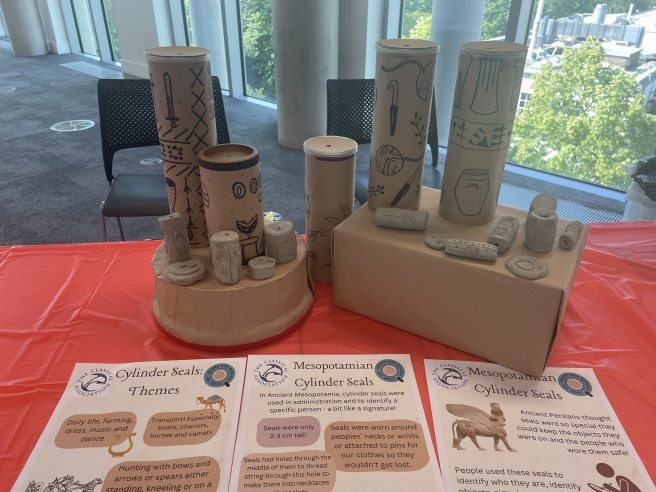 Small clay cylinder seals and large cardboard models of cylinder seals displayed on a table with a red table cloth.