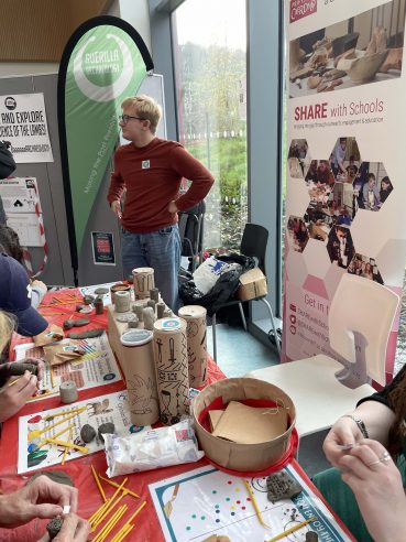 A busy table with examples of cylinder seals and hands around the table making their own. A banner stands behind the table with someone dressed in a. red jumper and jeans talking to a visitor off camera in the background.