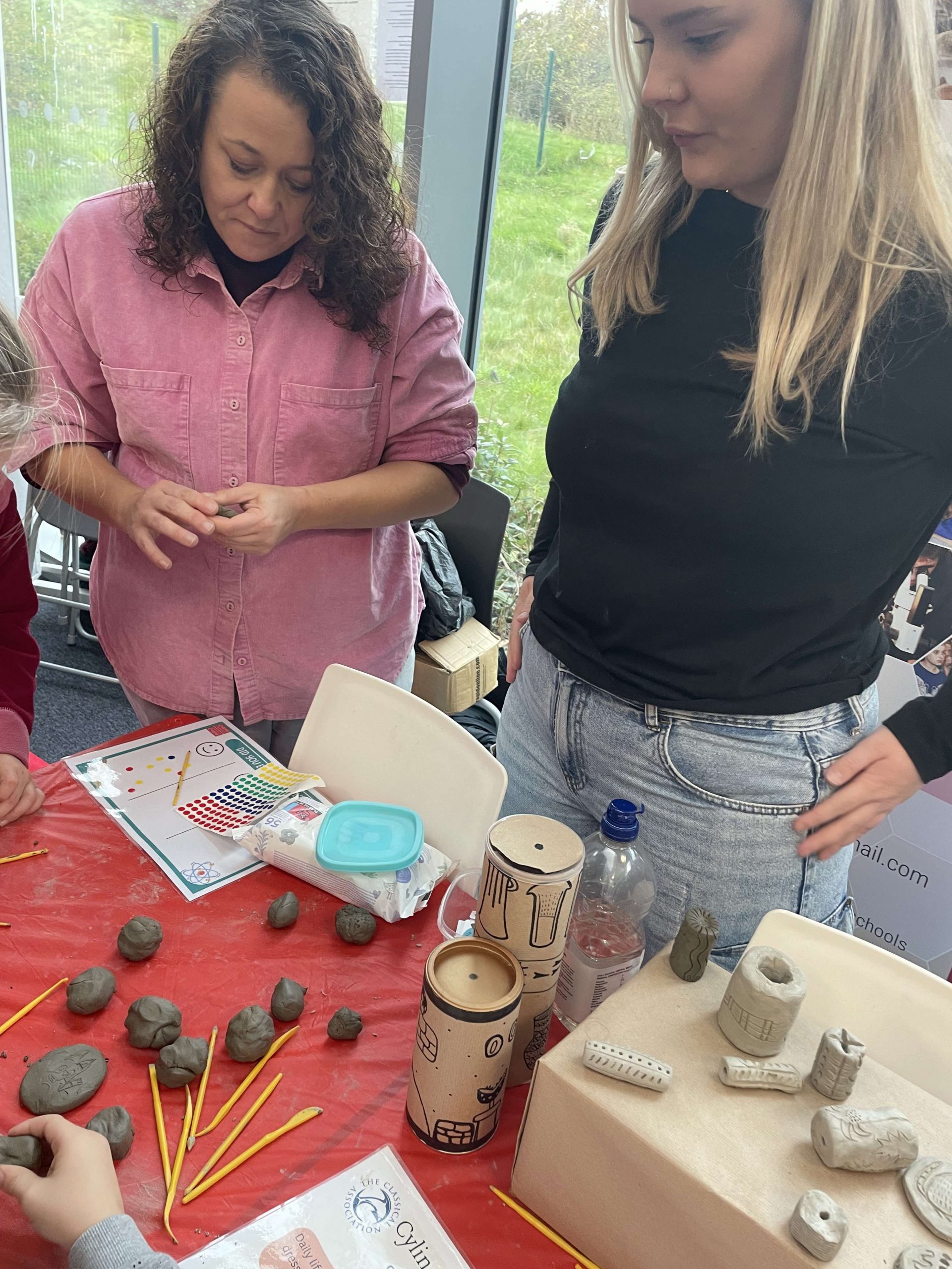 @people stand around a table making cylinder seals out of clay and talking to others off camera.