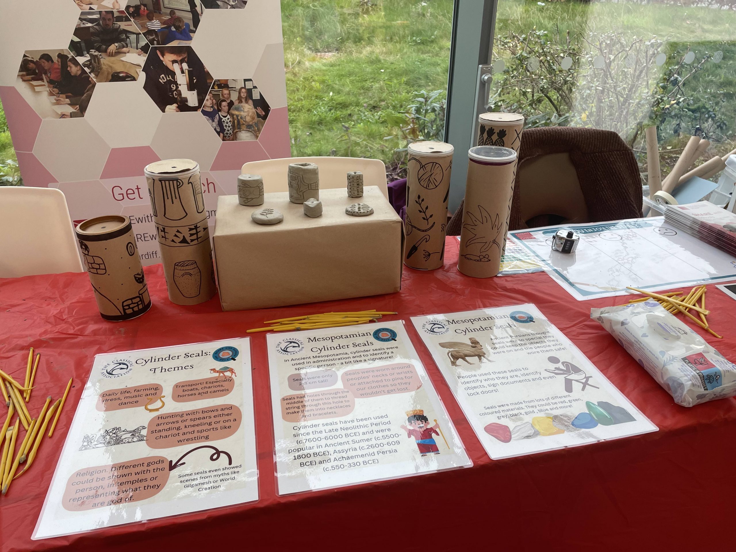 Model cylinder seals out of clay stand on a brown box with large scale seals made of crisp cartions either side. The table is covered in a red tablecloth with 3 info sheets infant of the seals.