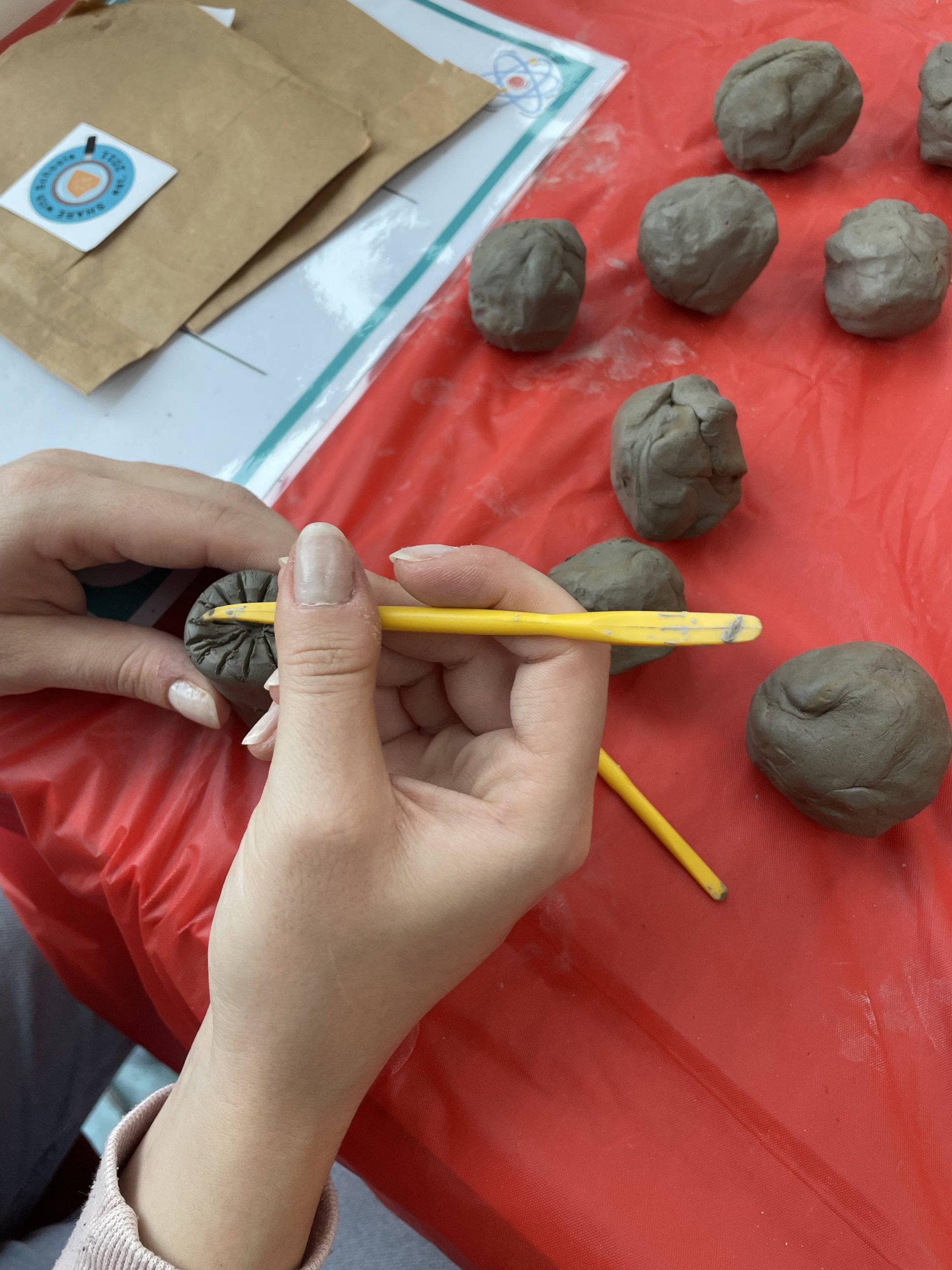 Balls of clay on a red table cloth. Over the table a pari of hands make their own seal with clay tools.